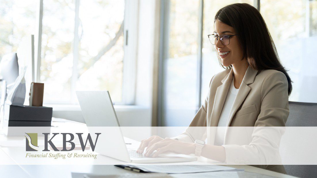 woman with long dark hair and glasses wearing a beige blazer works on her laptop in front of an office window